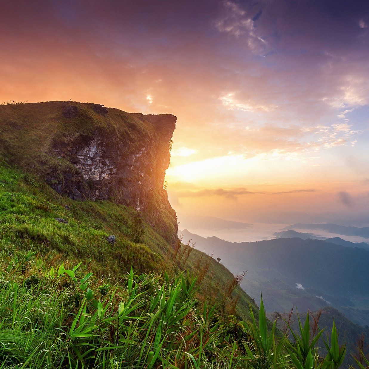 Breathtaking view of a mountain cliff at sunrise with vibrant skies and a lush green valley below.