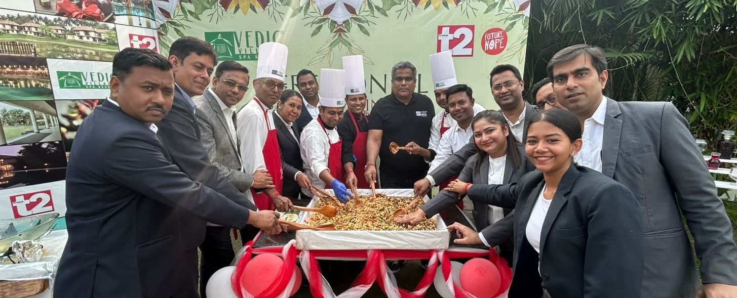 A group of people in formal attire are gathered outdoors, cutting a large cake on a decorated table with balloons | Vedic Village Spa Resort, Kolkata