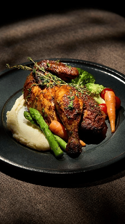 Plate of roasted chicken served on a bed of mashed potatoes with asparagus, carrots, and broccoli, dramatically lit against a dark background.