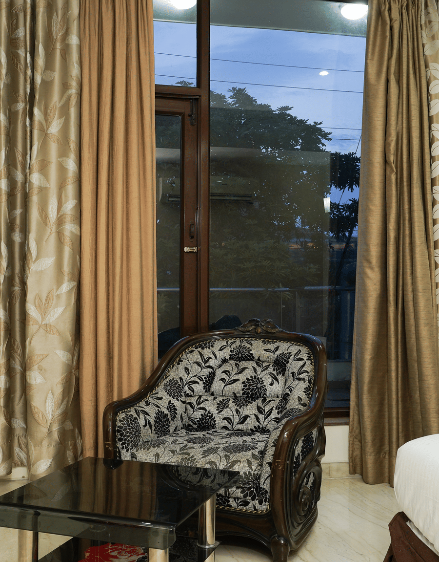 Carved armchair beside full-length curtains and a glass balcony door in the Deluxe Room at Lime Tree Hotel, IFFCO Chowk