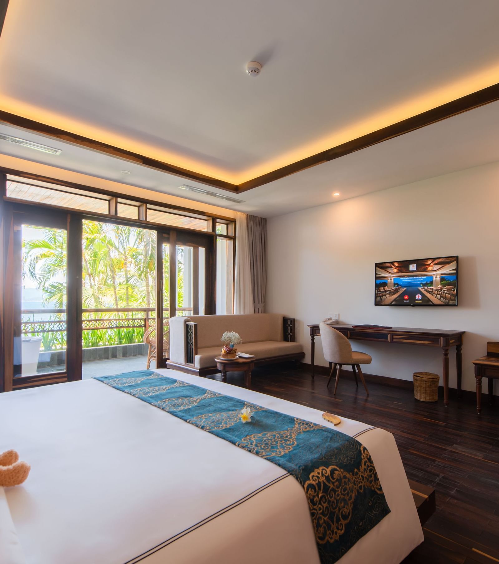 A hotel room with a large bed facing a television, furniture to relax and a balcony with a view of the greenery - Alibu Resort Nha Trang