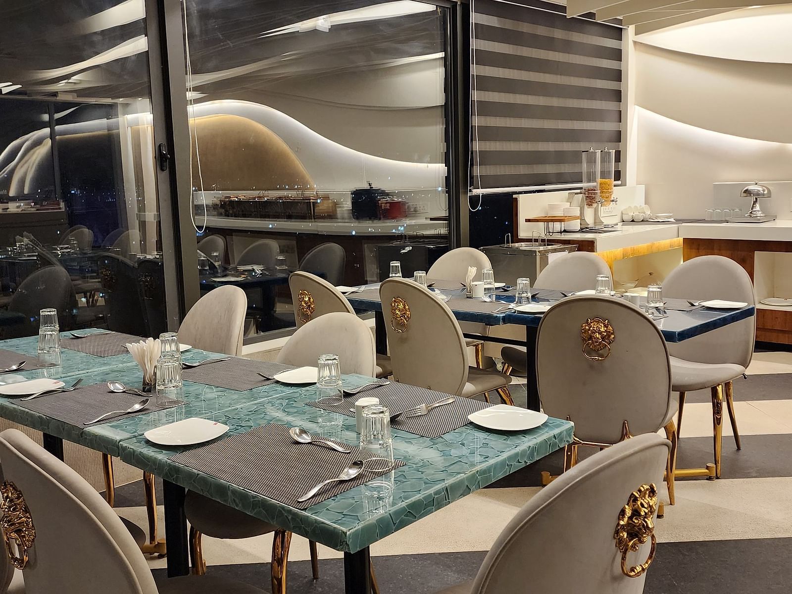 An elegant dining area with white and gold chairs and set tables, located beside large windows offering a view outside.