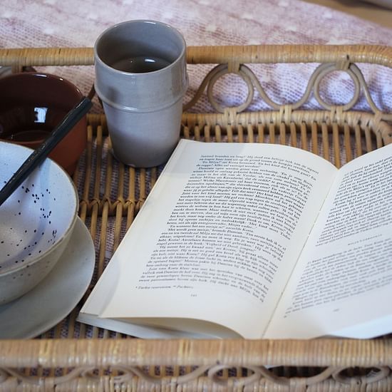 a book next to a bowl and a mug that is placed on a tray on a bed