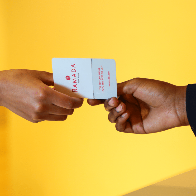 A close up of a two people exchanging a ramada visiting card with a yellow wall in the background.