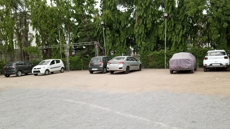 An outdoor open-air gravel parking lot with several cars parked in front of a line of green trees.