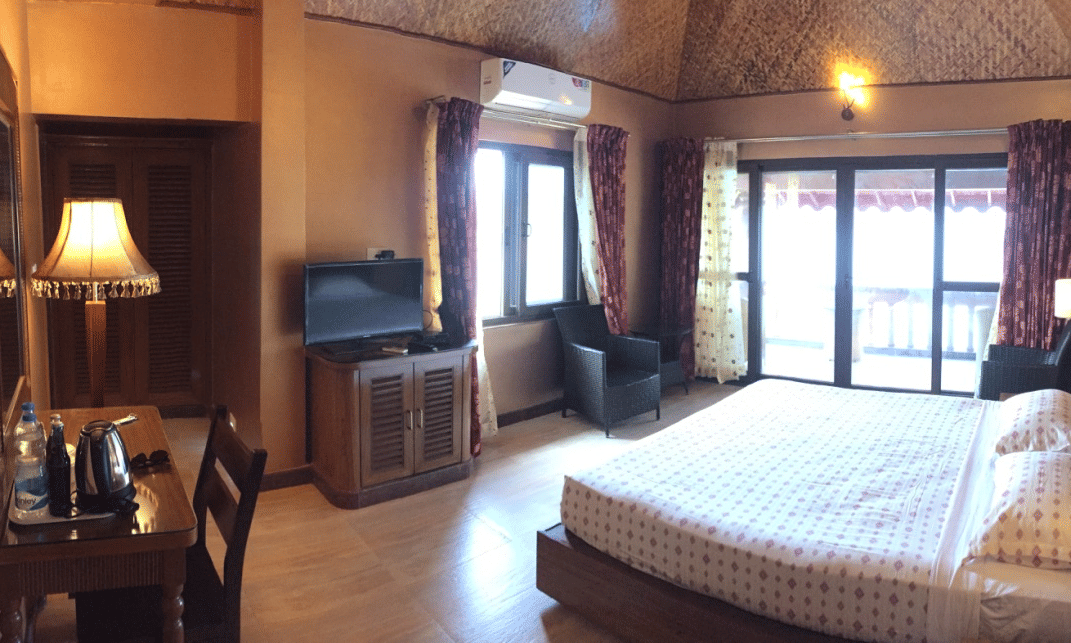Spacious bedroom with queen size bed, wooden flooring, side lamps, and large windows with sheer curtains at Paradise Lagoon Resort, Udupi.