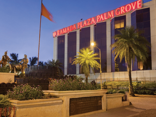 Exterior view of Ramada Plaza by Wyndham Palm Grove, showcasing its illuminated facade, palm-lined surroundings, and prominent city landmark setting.