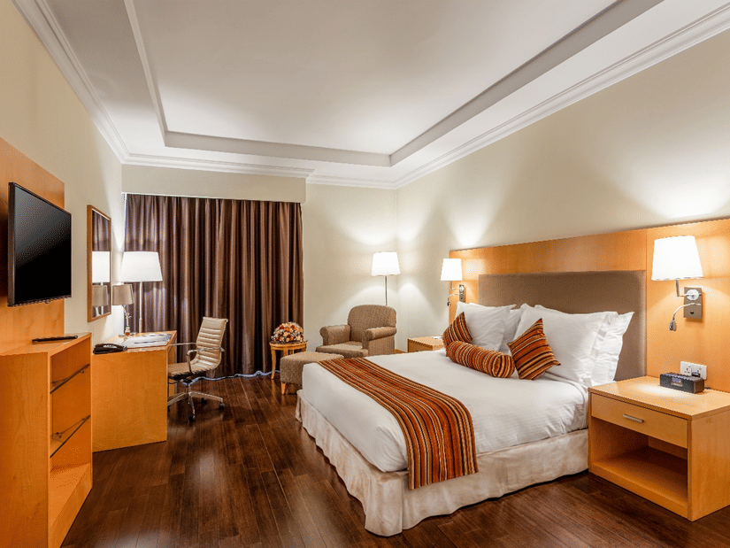 A modern hotel room at Ramada Plaza by Wyndham Palm Grove, designed with warm interiors, a plush bed, a work desk, and a wall-mounted television.