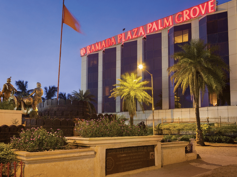 Exterior view of Ramada Plaza by Wyndham Palm Grove, showcasing its illuminated facade, palm-lined surroundings, and prominent city landmark setting.
