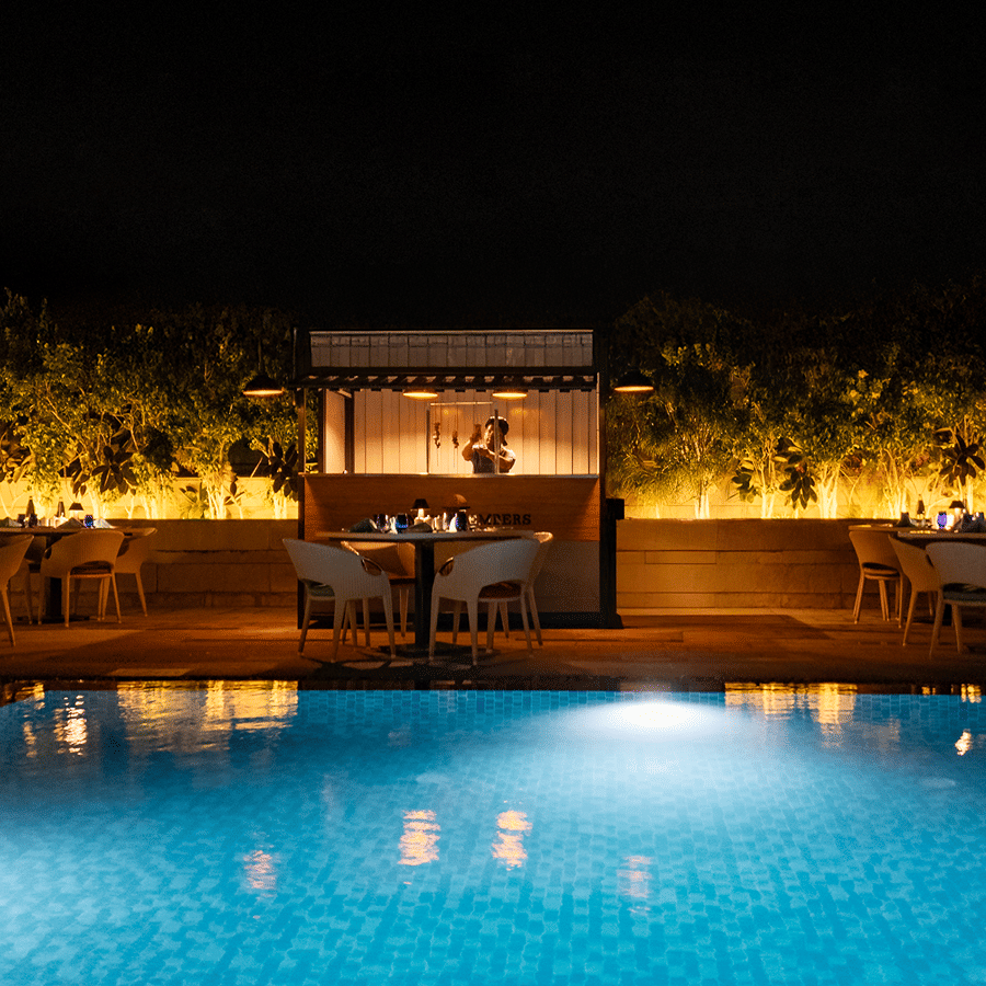 A tranquil nighttime view of the hotel's outdoor swimming pool, lit up with glowing lights, with a seating area surrounded by plants at The Residency Towers Rameswaram.