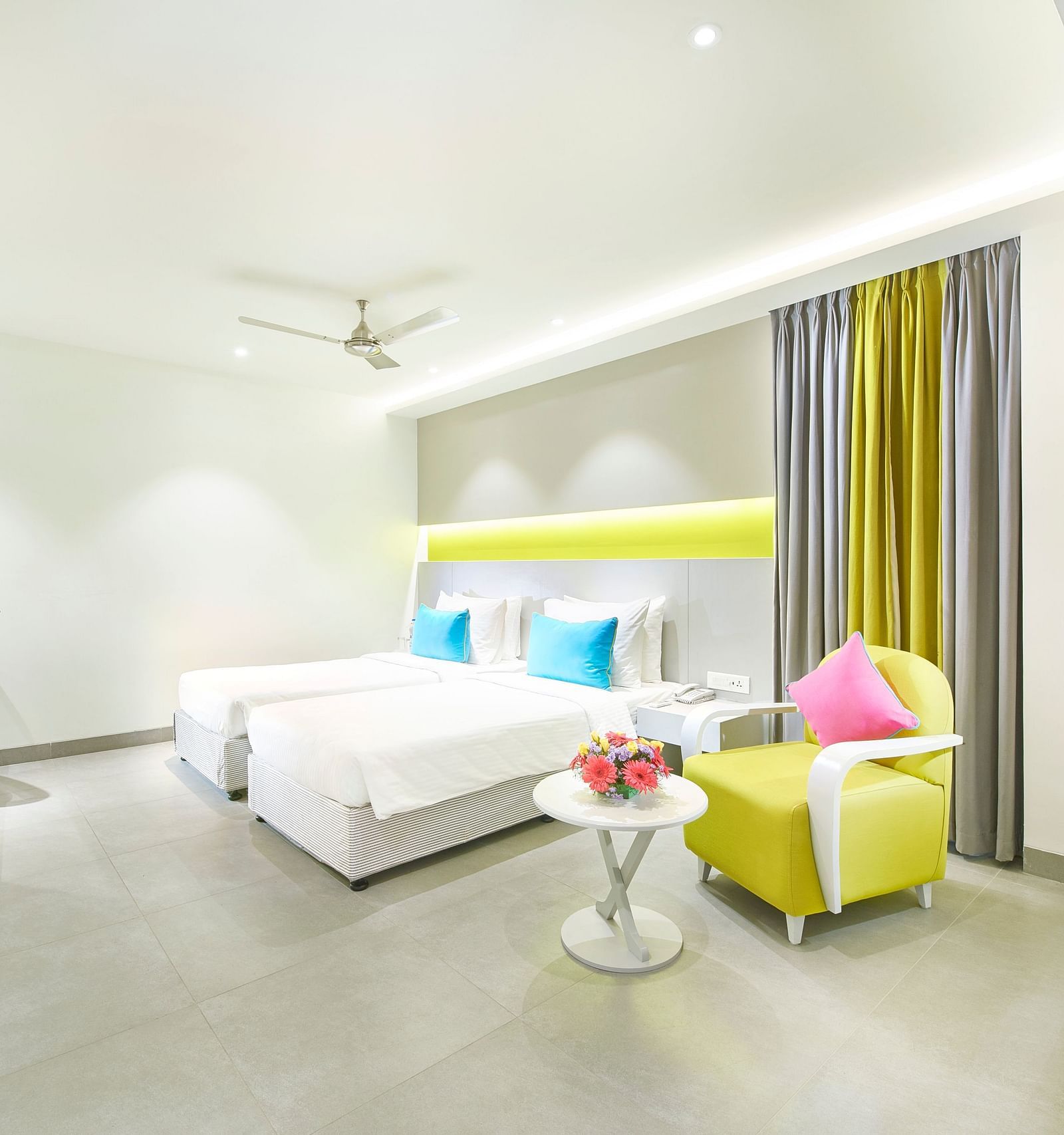 A spacious room features a large bed with a headboard stretching across the wall. Cushion chairs sit beside a wooden bedside table, and a wall-mounted television is positioned opposite the bed