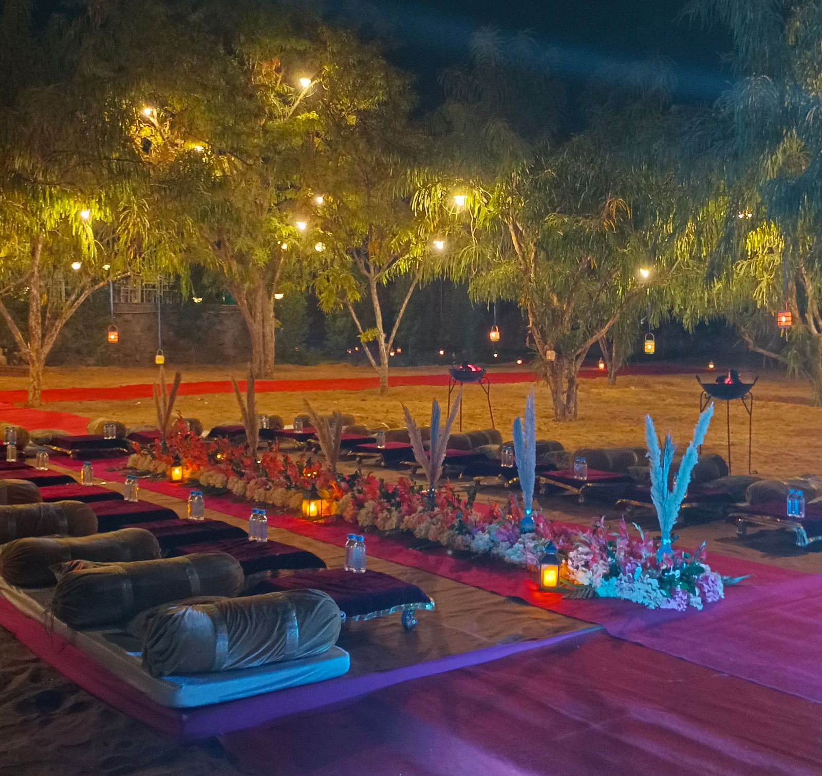 An outdoor area at night at the Bamboo Saa Mulberry Resort, Pushkar, with seating arrangement on the ground illuminated by string lights.