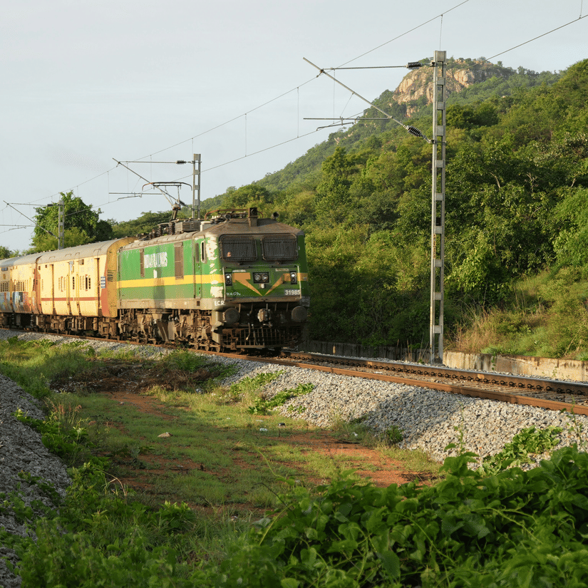 A long passenger train with green and yellow coaches travels along a curved track through a lush green landscape with a large hill in the background.