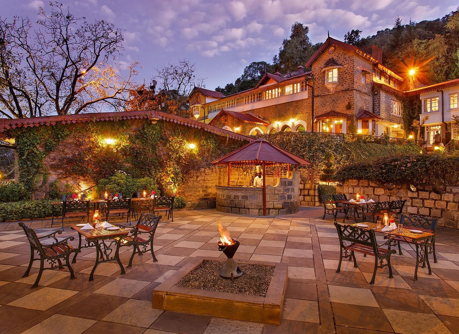 A beautiful outdoor dining area at the naini retreat, nainital. There are tables and chairs set up on a stone patio, with a fireplace in the center. The building is lit up at dusk, creating a warm atmosphere. 