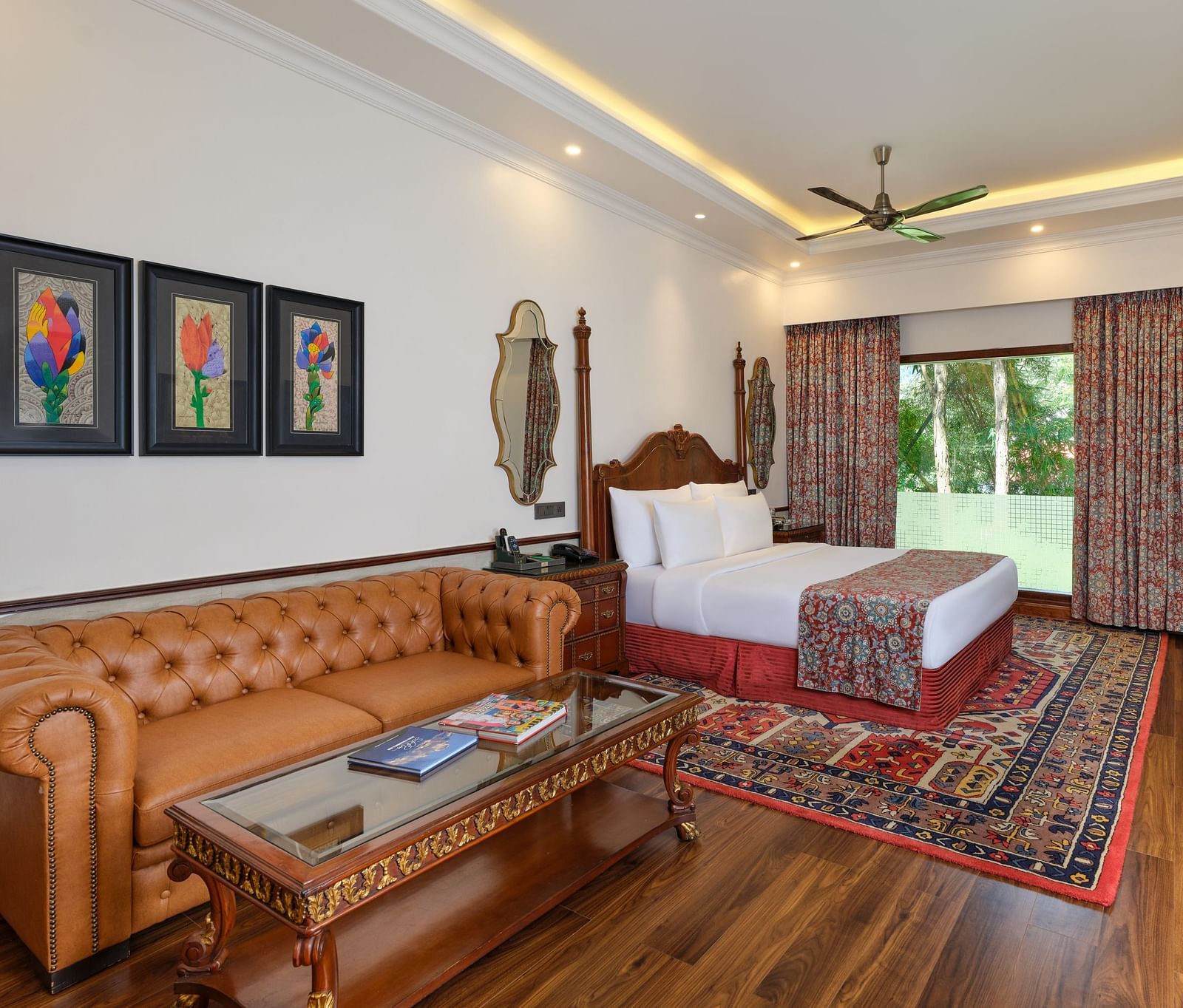 a Crown Villa bedroom with a large bed on a decorative rug, sofa, wooden cabinet, TV, artworks, and a window overlooking greenery  