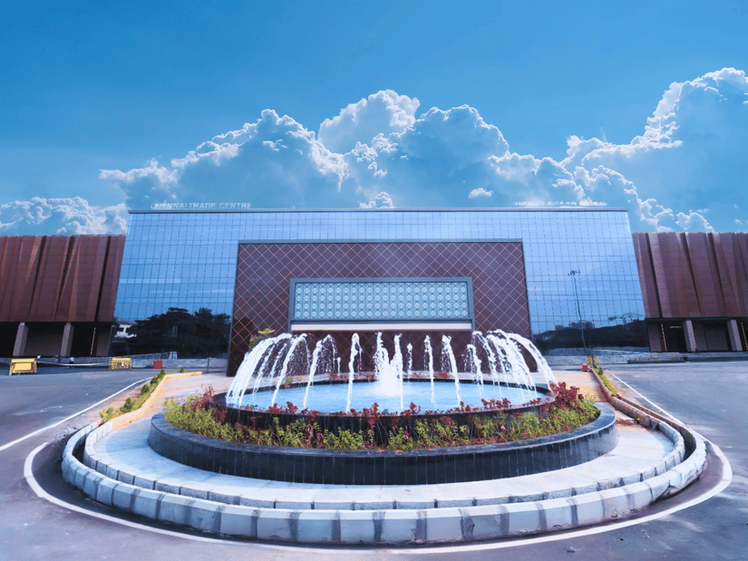 The modern, reflective glass facade of the Chennai Trade Centre, with a central fountain and a dramatic blue sky with white clouds overhead.