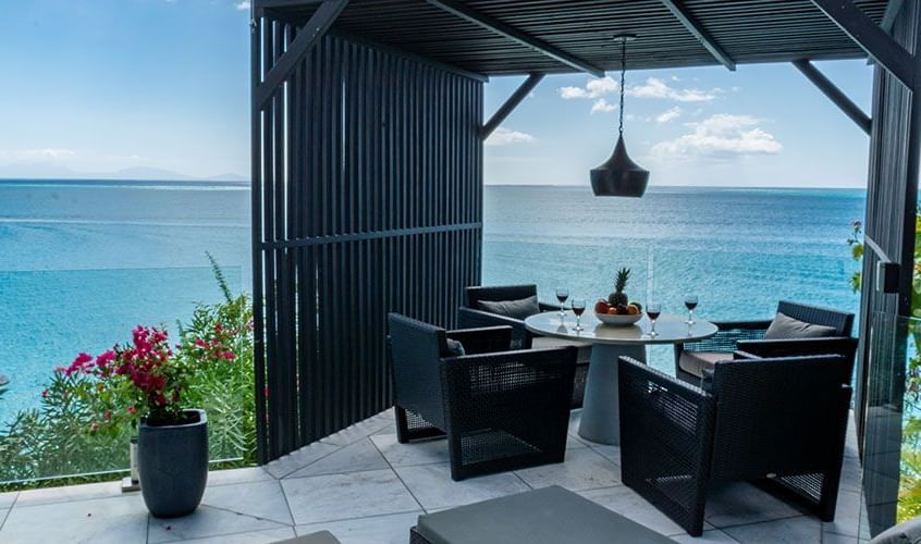 An elegant outdoor dining area with dark wicker chairs and a round table set with fruit and wine glasses, located under a modern pergola with a panoramic view of the blue ocean.