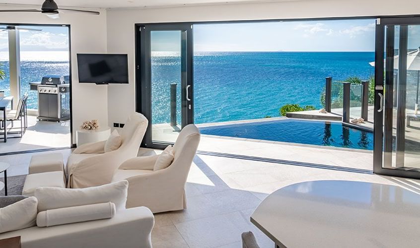 A bright, modern living room with white furniture opens up to an expansive ocean view through large glass doors, leading to an outdoor deck and turquoise water - Tamarind Hills Resort and Villas - Antigua