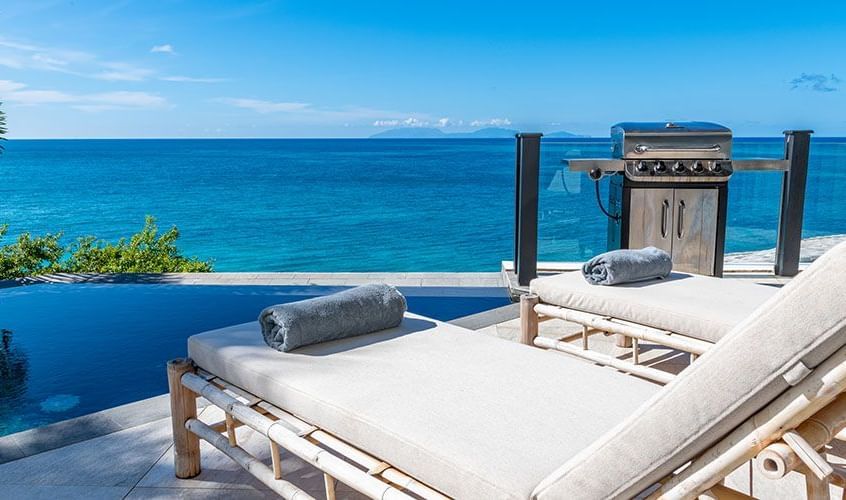 Rolled towels placed on sunloungers and an outdoor grill that are beside the deep blue infinity edge pools that seamlessly blends into the Caribbean Sea beyond Tamarind Hills Resort.