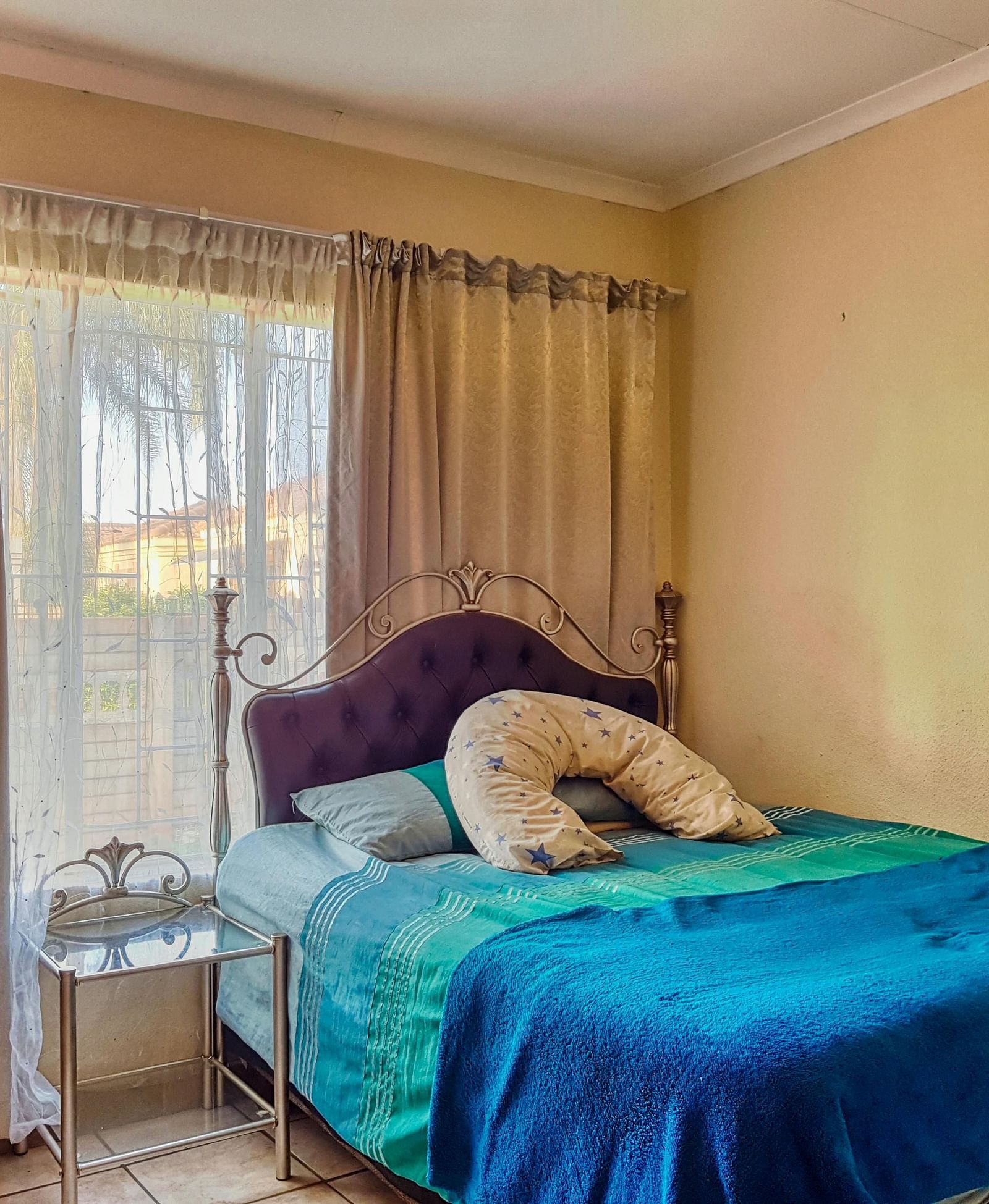 A bedroom featuring a double bed with blue sheets spread neatly over it with beige curtains covering the window behind it.