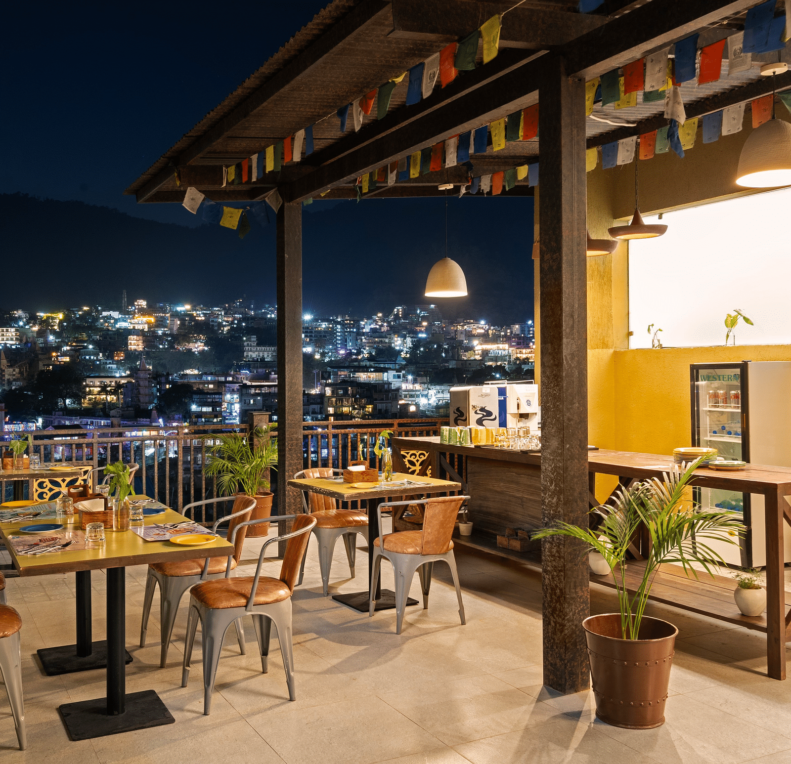 Panoramic view of city skyline from rooftop restaurant, with tables, chairs, and city lights at night at Bedzzz, Rishikesh 