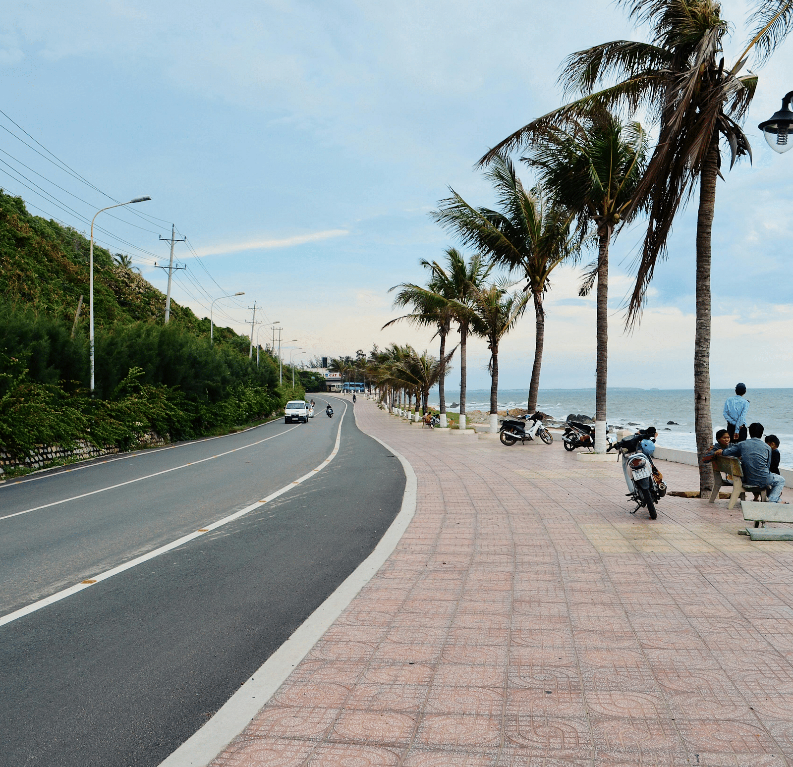 A coastal road with palm trees lining a pedestrian walkway and people sitting on benches overlooking the ocean.