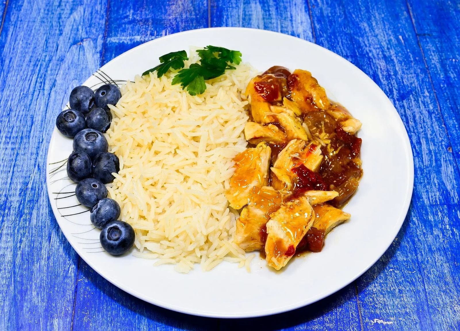 A white plate holds a serving of rice, with meat, and blueberries, presented on a blue textured surface | Altamont Court, Jamaica