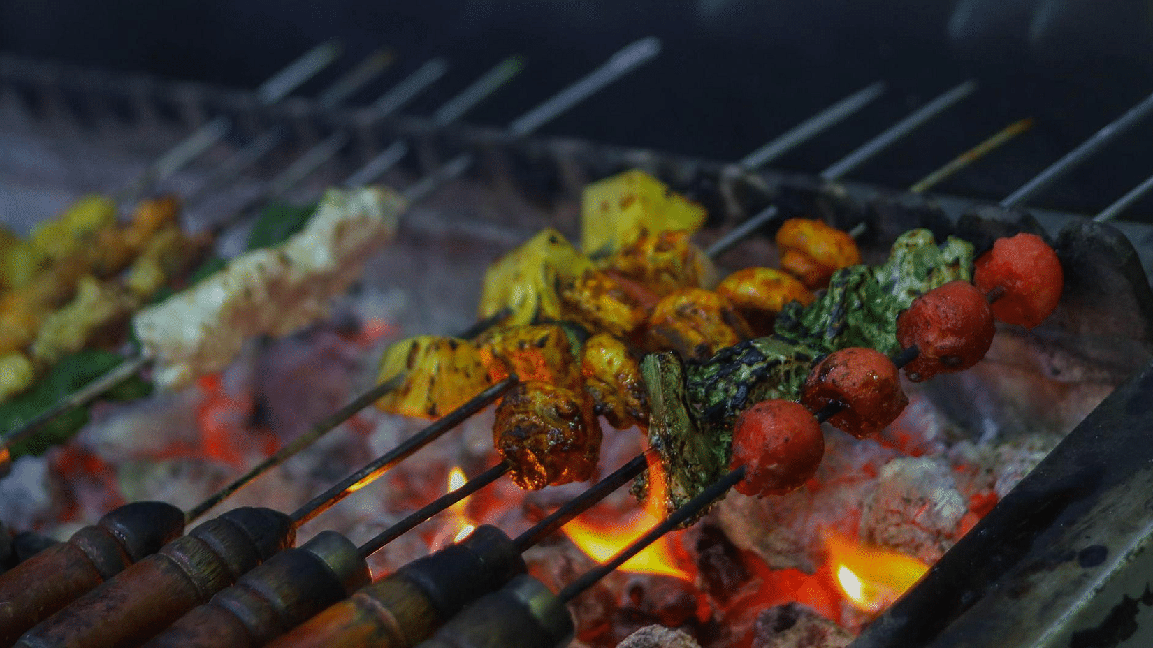 Skewers of colorful marinated vegetables and paneer grilling over hot coals with visible flames - Jenneys Residency, Coimbatore