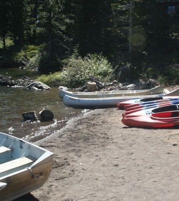 Several canoes and kayaks are beached on the sandy shore of a lake surrounded by trees.