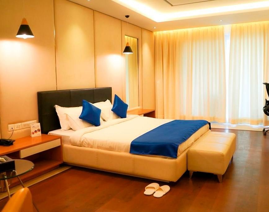 A warm and inviting hotel room at BNGV Grandeur Hotel and Banquets, featuring a bed with blue linens, wooden accents, and soft ambient lighting.