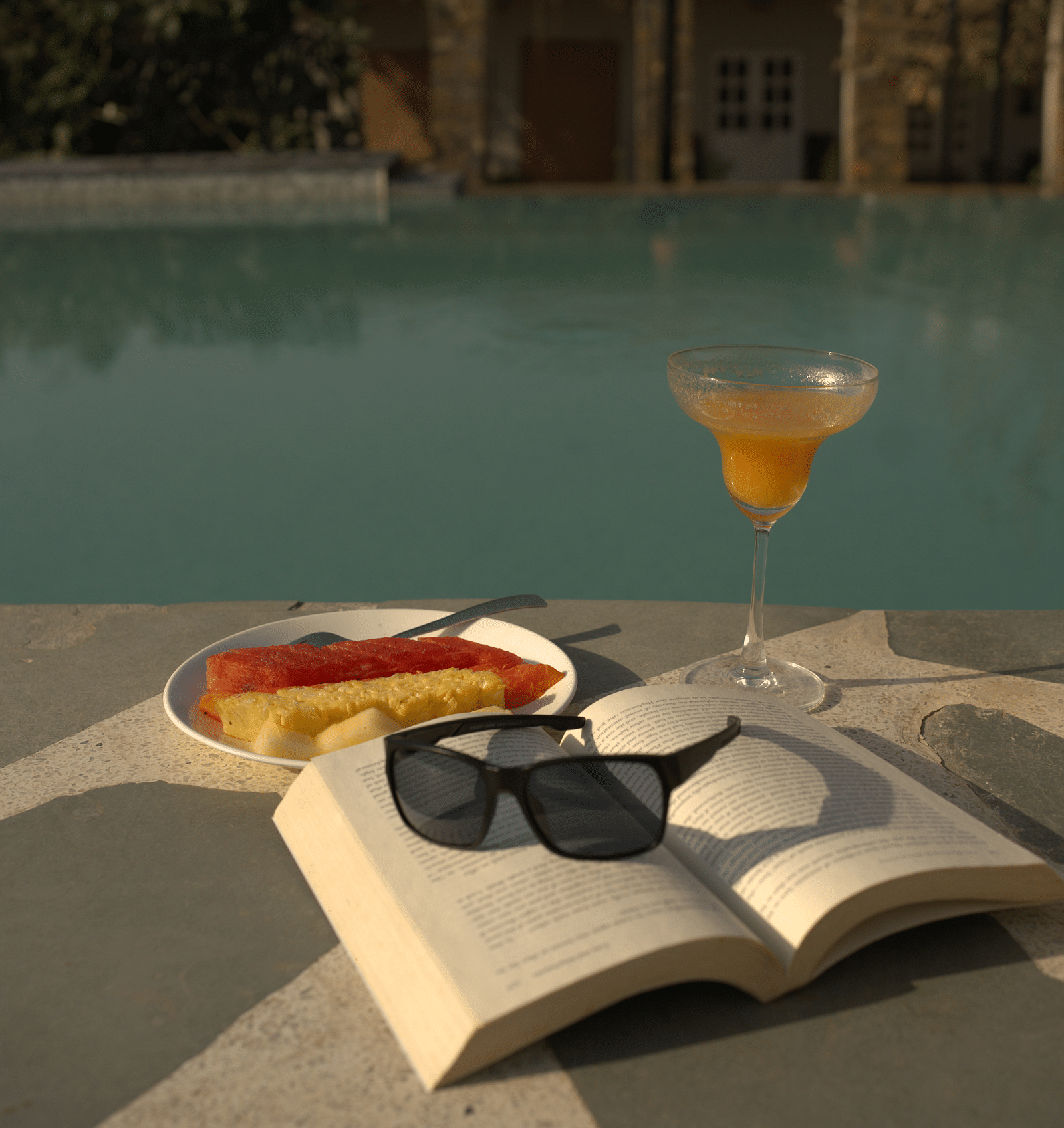 An image of a book, sunglass, breakfast and beverage near a pool
