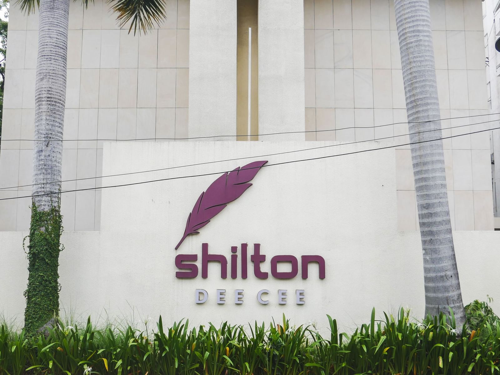 The exterior of Shilton Dee Cee, showing the hotel's prominent purple logo and lush green landscaping.