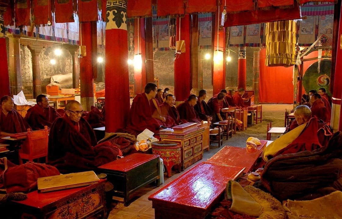 An image of a well lit monastery with lamps hanging from the ceiling and below it monks have gathered together