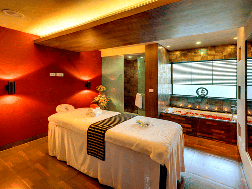 An overview of Sparoh Spa in Kolkata with a massage bed, a bathtub filled with water and rose petals, tea light candles next to the tub and aesthetic lighting at Kenilworth Hotel, Kolkata.