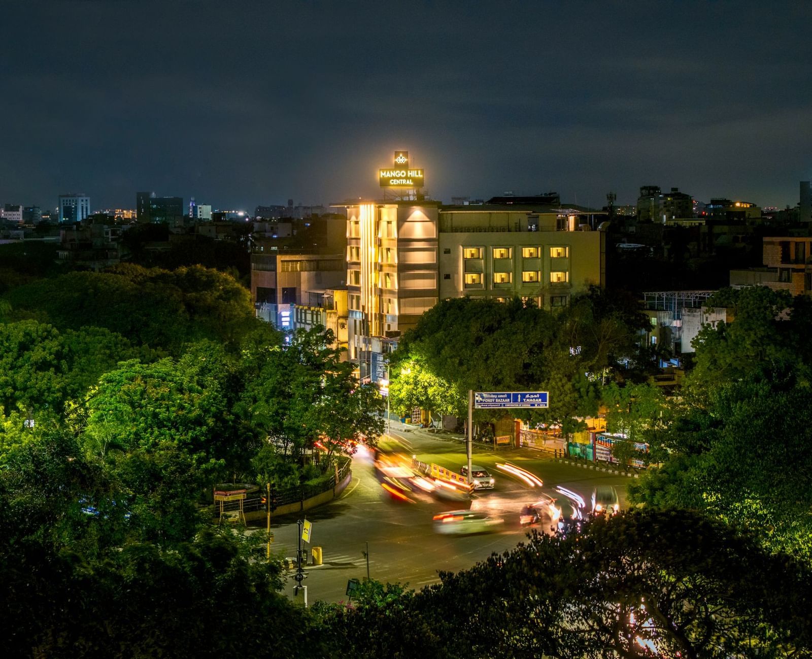 An aerial night view of Mango Hill Central, Chennai, showing the illuminated hotel building and outdoor seating area.