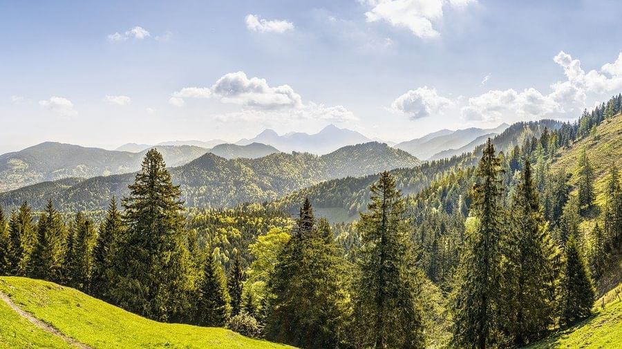 A panoramic picture of a hilly area with a house, trees and mountains in view.