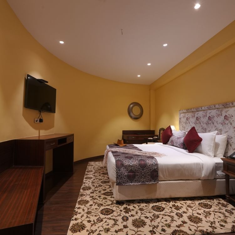 Side view of the double bed, a wall mounted television and a wide table in the Suite Cottage Room at The Garden Ananta Elite Rajkot 4