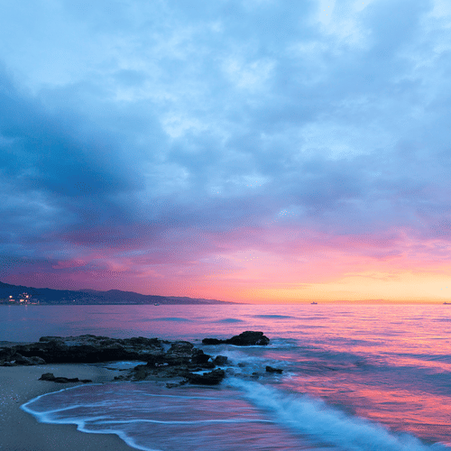 Purple and pink sunset over a rocky beach with gentle waves and a distant city skyline under a cloudy blue sky.