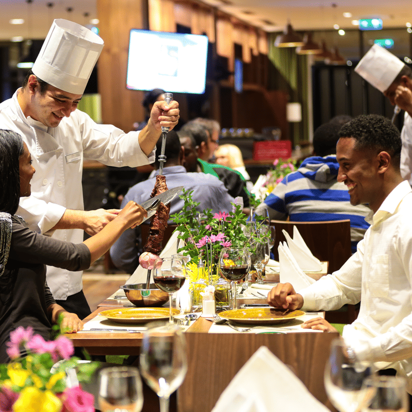 A couple sitting inside a restaurant while the chef is serving them with other people in the background - Ramada by Wyndham Addis, Addis Ababa.