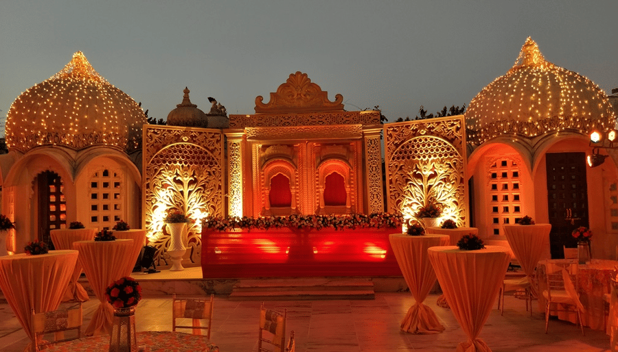An ornate wedding stage and reception backdrop setup at Pratap Chowk, flanked by traditional palace domes decorated with golden fairy lights at Chunda Palace, Udaipur.