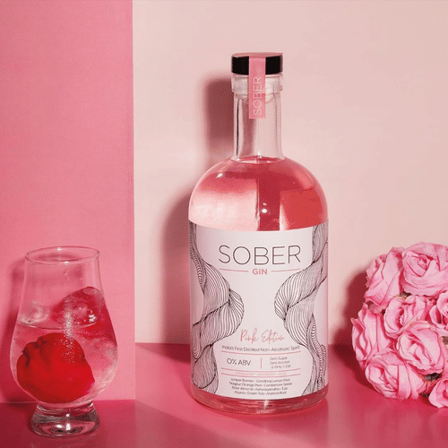 A bottle of Sober Gin placed beside a bouquet of pink roses and a wine glass containing a pink fruit against a pink backdrop