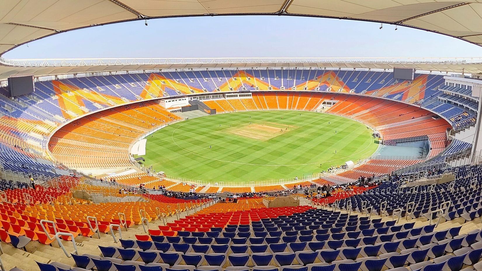 Wide-angle view of Narendra Modi Stadium with green cricket field, orange and yellow seating sections, and a partially covered roof.