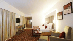 A well lit room featuring a neatly arranged bed, a sofa, a wall-mounted TV, and photo frames on the wall