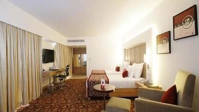 A well lit room featuring a neatly arranged bed, a sofa, a wall-mounted TV, and photo frames on the wall