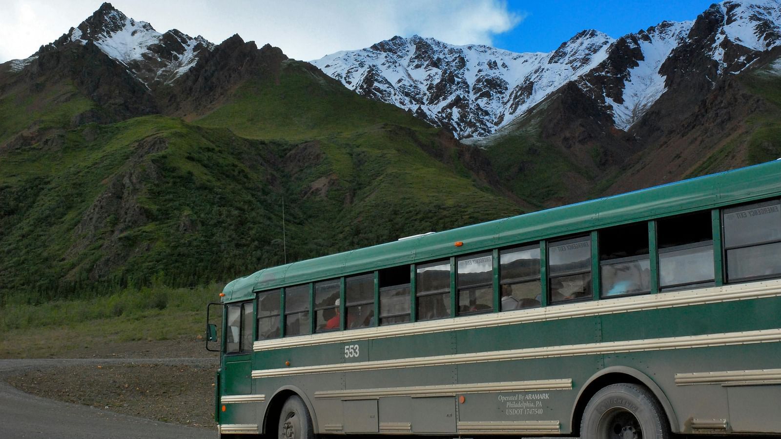 Green and white bus travelling on a road with snow-capped mountains in the background.
