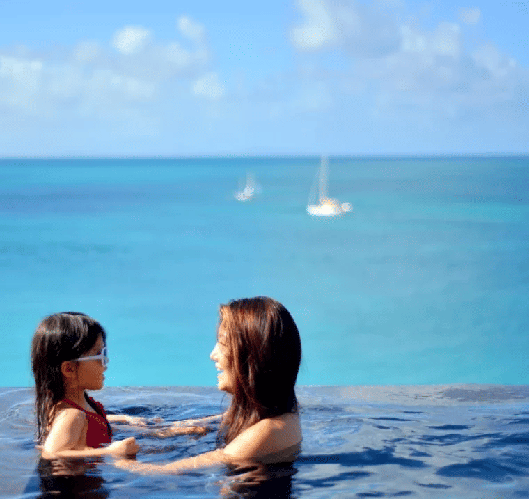 Two people in an infinity pool with a view of sailboats on the turquoise ocean