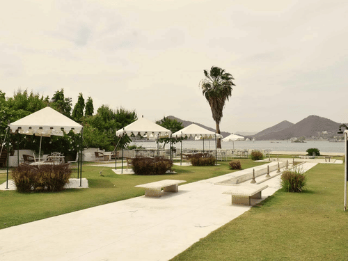 Outdoor seating area with white tents, benches, and lush green lawn by a lake with hills in the background at Ram Pratap Palace, Udaipur.