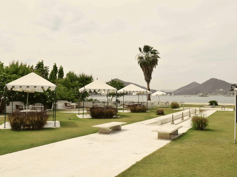 Outdoor seating area with white tents, benches, and lush green lawn by a lake with hills and Fateh Sagar Lake in the background at Ram Pratap Palace, Udaipur.
