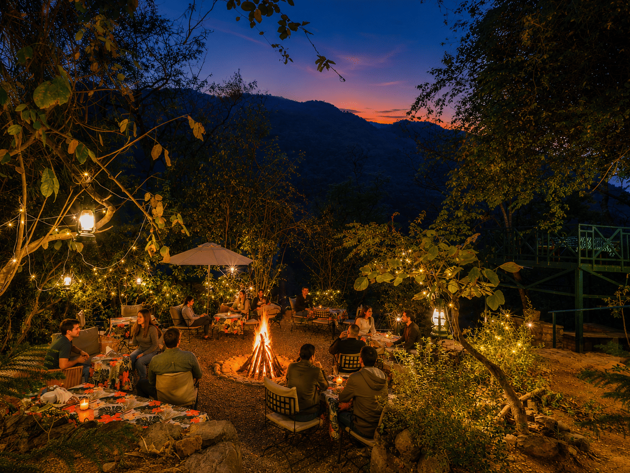 Cosy evening bonfire at Neemrana’s Glasshouse on The Ganges, Rishikesh, with guests relaxing outdoors amidst trees against a sunset over mountains.