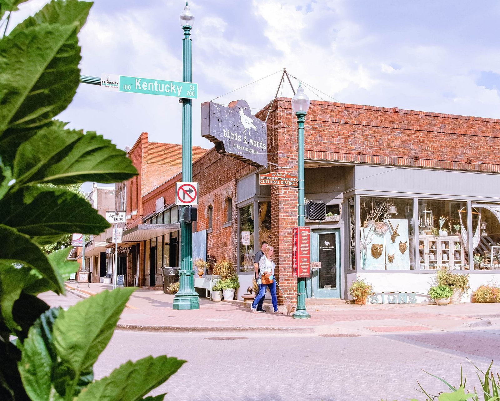 A crossing with a marketplace with brick like textures on the exterior with physical signs to show directions, a couple walking on the road and green leafs on the side.